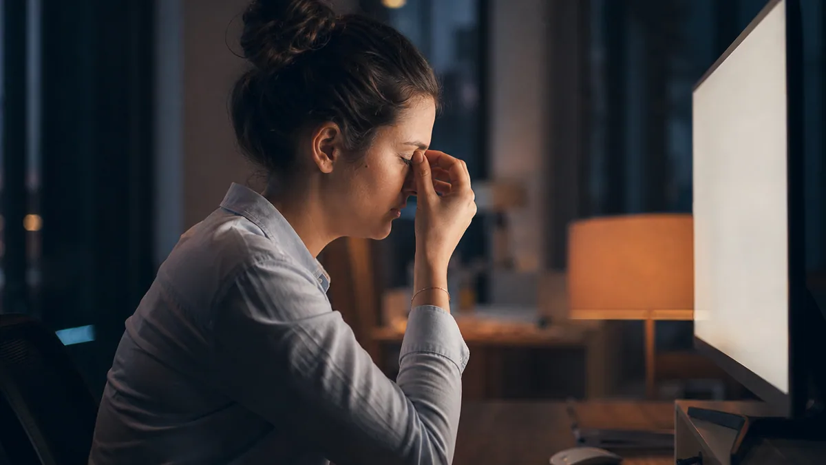 Personne se frottant les yeux devant un écran d'ordinateur en soirée, lumière tamisée